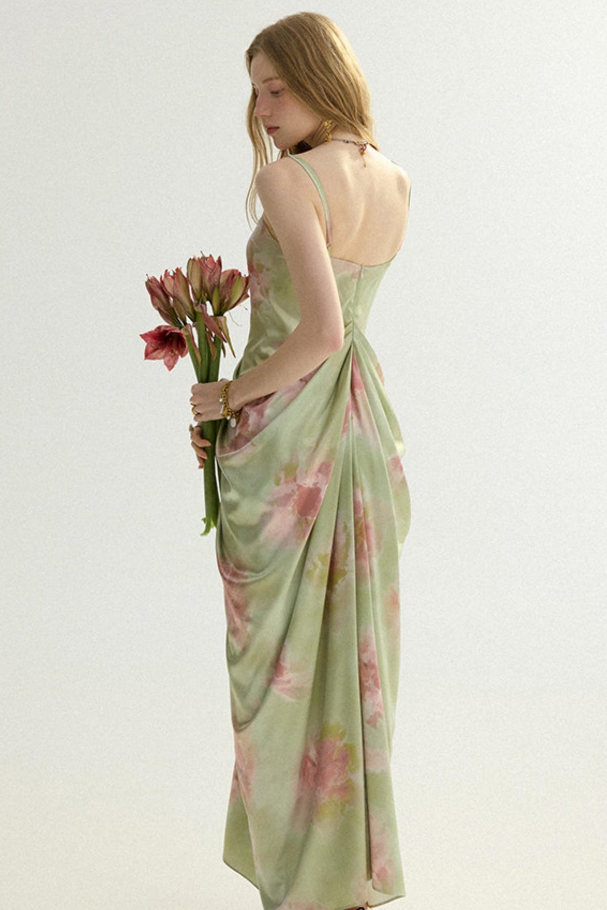 A woman wearing a light sage green satin maxi dress with a pink floral print, shown from the back while holding a bouquet of pink flowers against a plain light background.