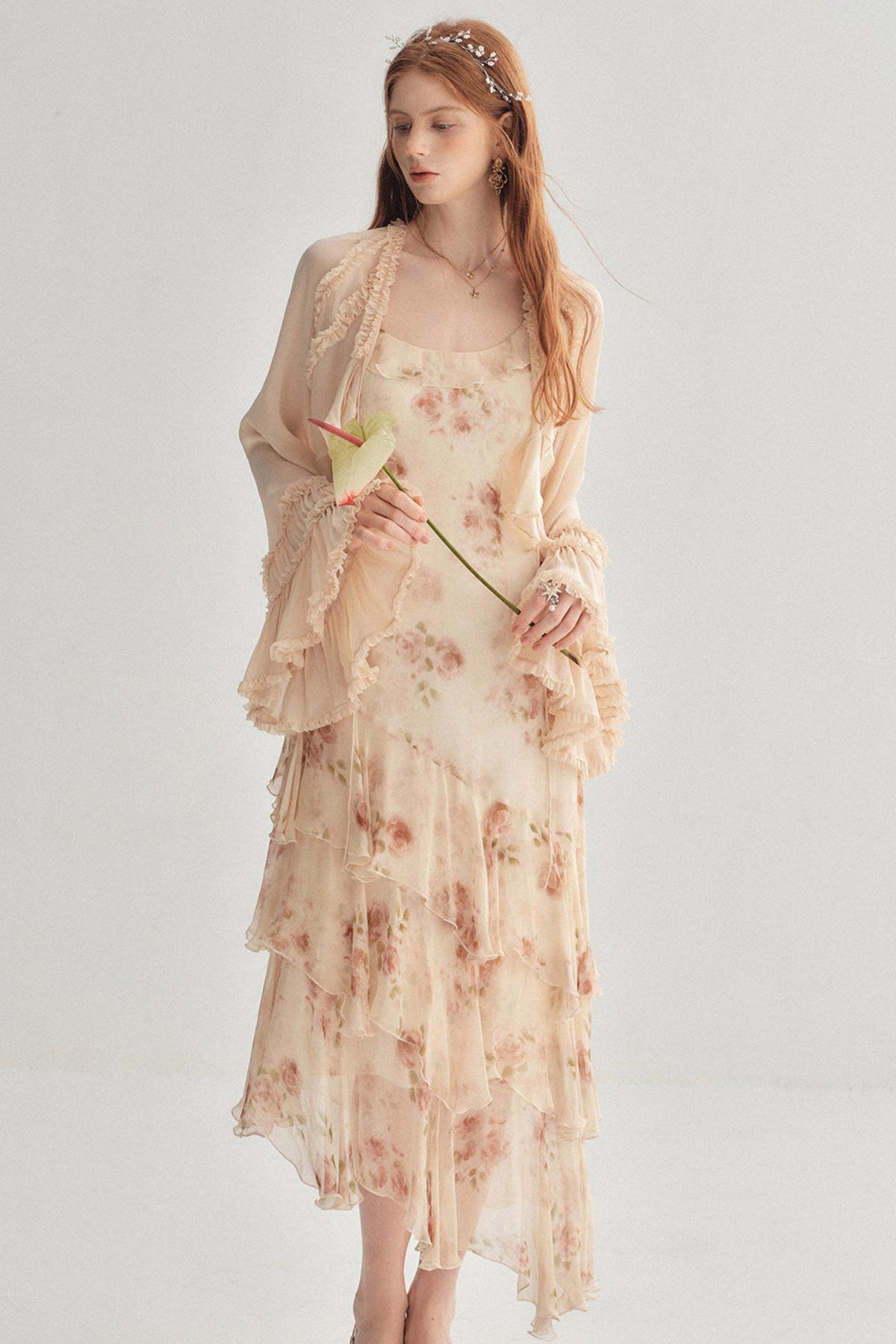 A woman modeling a sheer, long-sleeved beige dress with a soft pink floral print and a matching lace-trimmed cardigan, set against a plain light gray studio background.