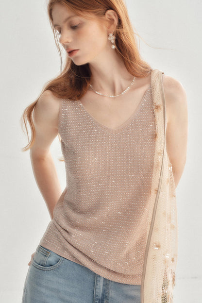 A woman wearing a beige sleeveless knit top covered in small shimmering rhinestones, paired with light blue jeans and a pearl necklace, against a clean white background.