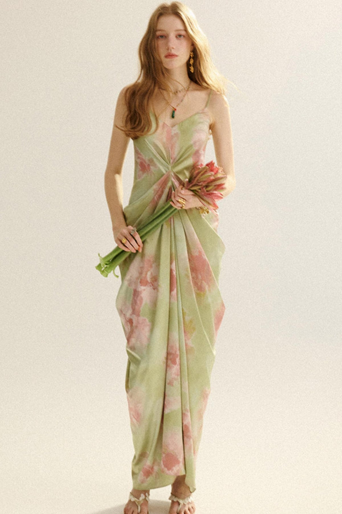 A woman modeling a light sage green and pink floral maxi dress with a twisted front detail, holding a bouquet of pink flowers against a plain beige background.