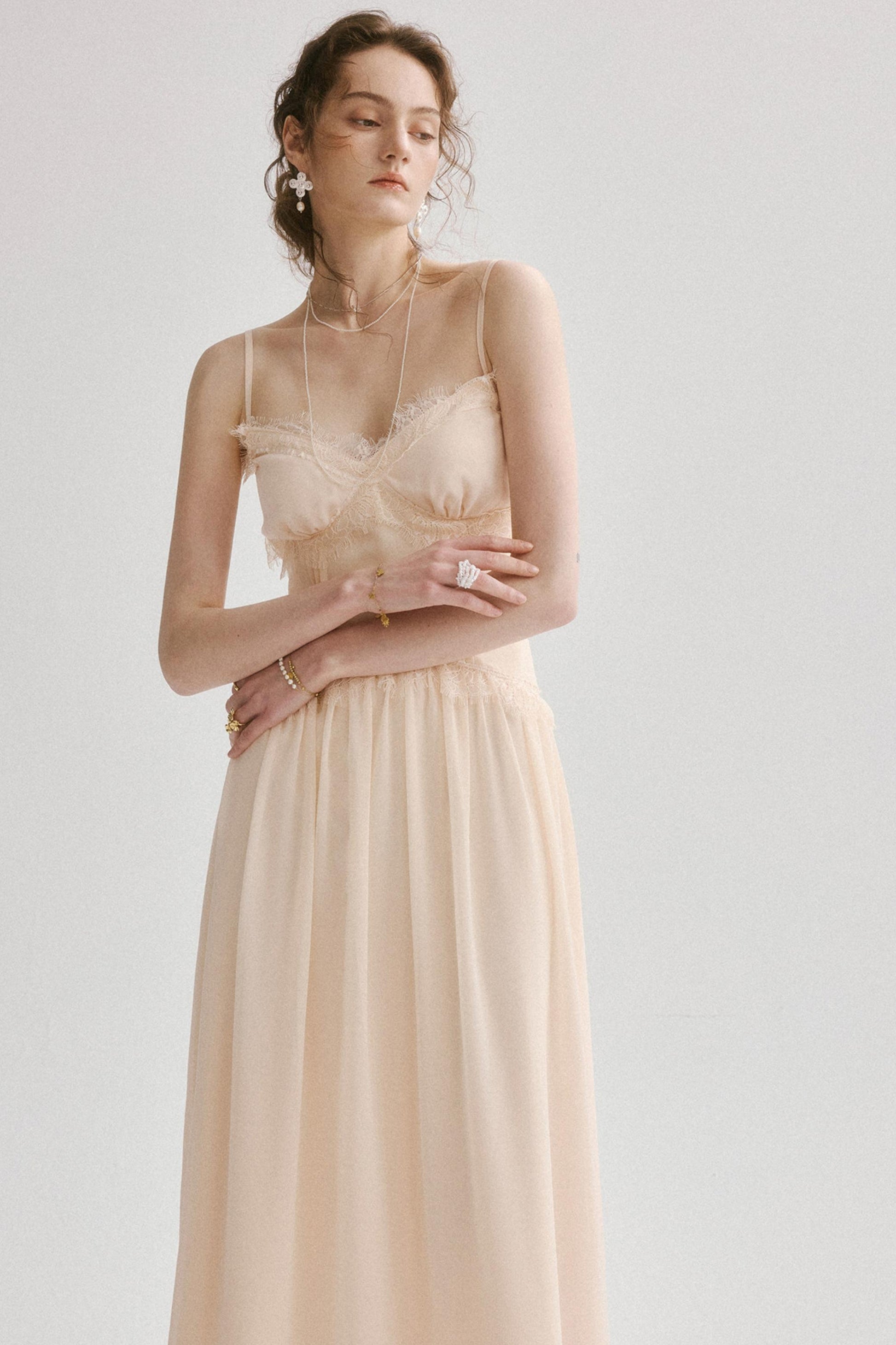 A woman modeling a light peach-beige maxi dress with a lace-trimmed sweetheart bodice and thin spaghetti straps, set against a plain light gray studio background.