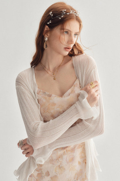 A woman wearing a sheer cream ribbed cardigan over a pale floral camisole dress, accessorized with a floral headband, gold necklaces, and rings, set against a plain light gray background.