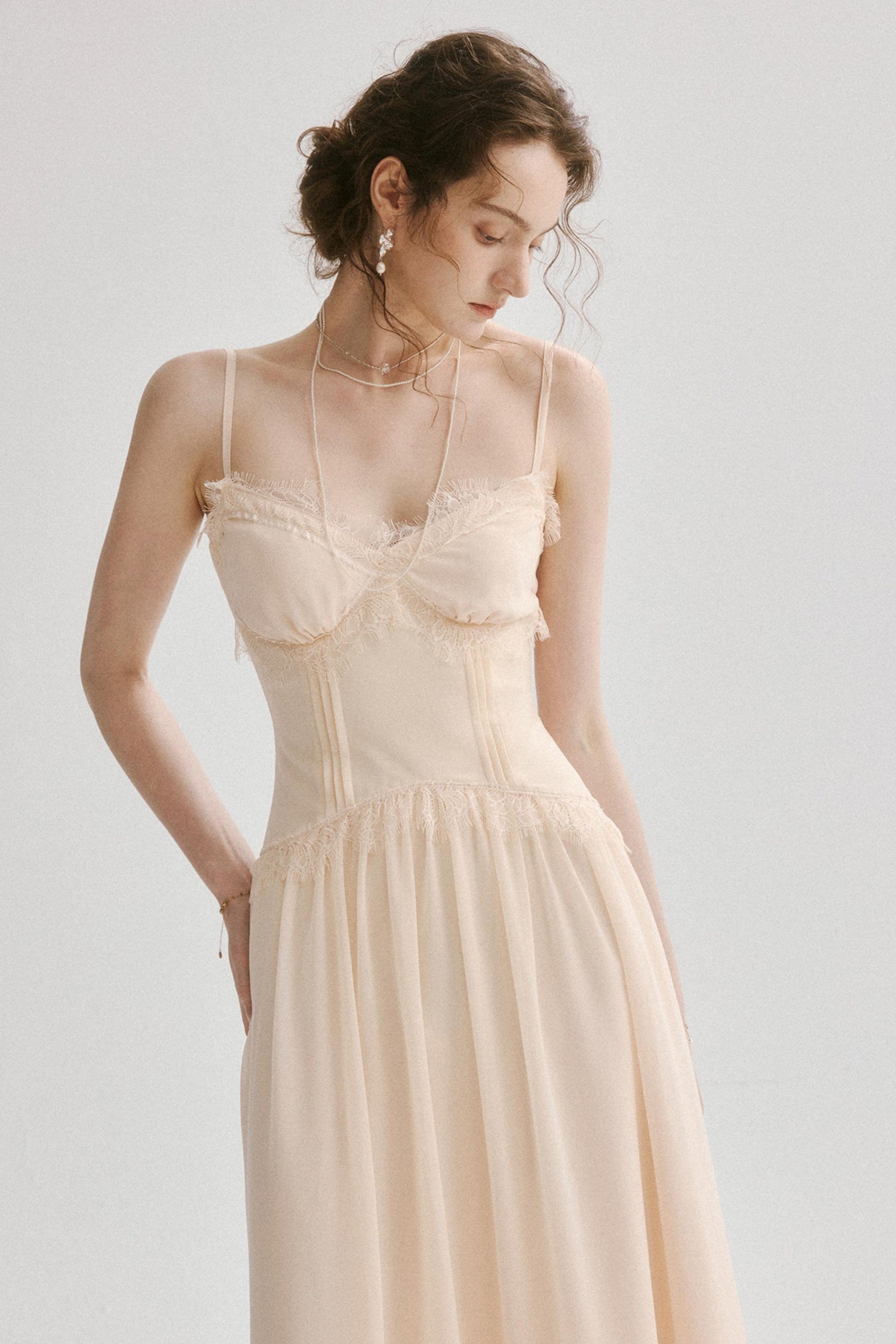 A woman wearing a light cream or pale beige sleeveless maxi dress with lace trim and a fitted corset-style bodice, photographed against a plain light gray studio background.