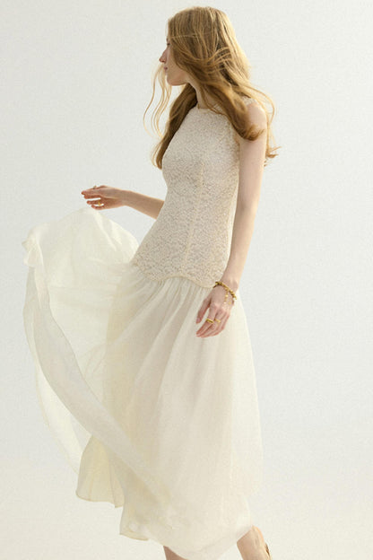 A woman wearing a sleeveless cream-colored dress with a textured lace bodice and a flowing chiffon skirt, captured in motion against a plain white studio background.