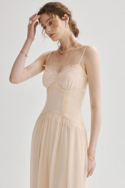 A woman wearing a light beige or cream-colored lace-trimmed camisole nightgown with a corset-style bodice and a flowing skirt, photographed against a plain light gray studio background.