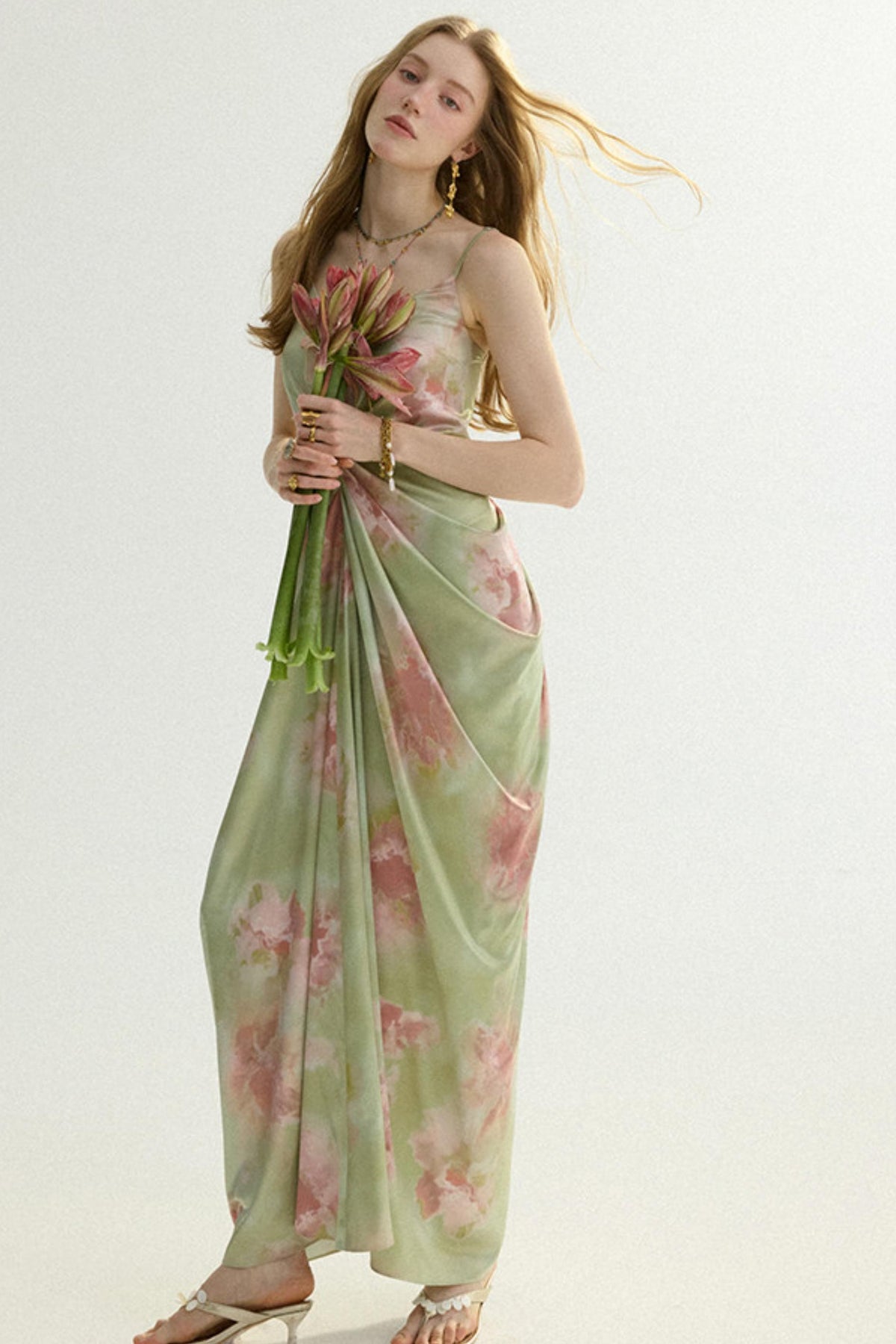 A woman modeling a light sage green maxi dress with a pink floral print, holding a bouquet of pink flowers against a plain white studio background.