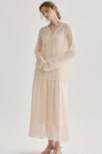 A woman modeling a cream-colored knit cardigan with lace-up front detailing and a matching sheer maxi skirt, set against a plain light gray studio background.