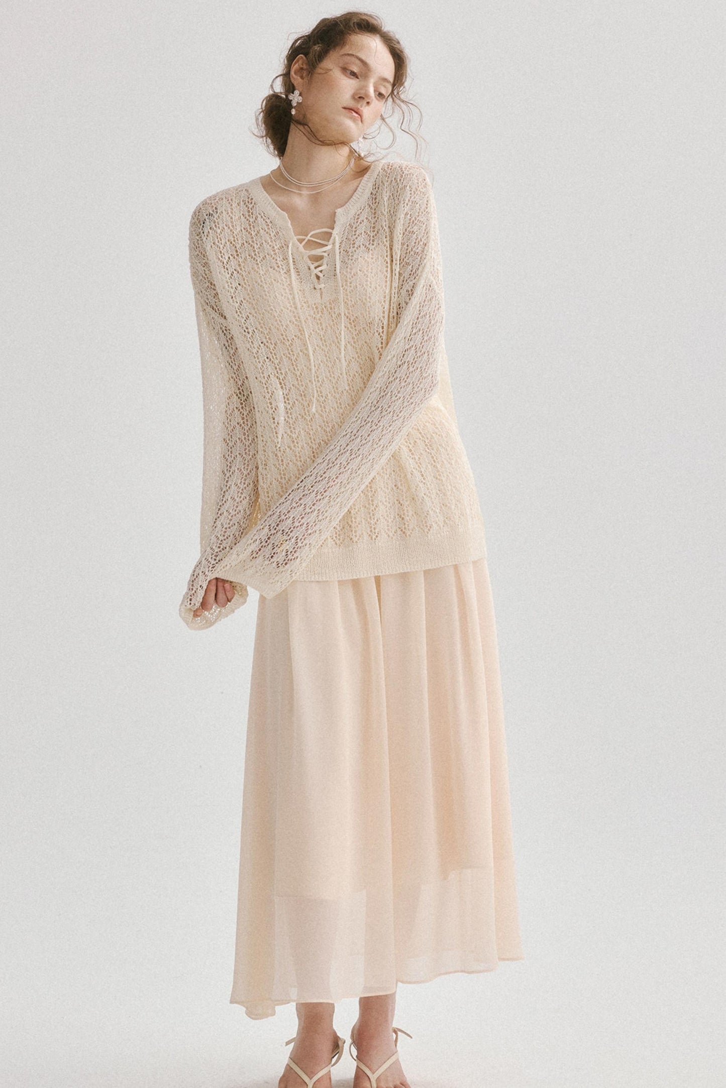 A woman modeling a light cream lace-up knit cardigan layered over a matching long-sleeve maxi skirt, set against a plain light gray studio background.