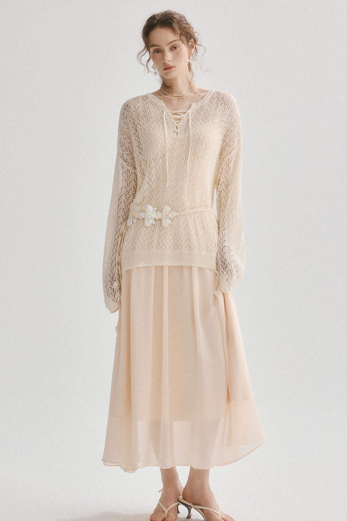A woman modeling a cream-colored two-piece outfit consisting of a long-sleeve lace top with a lace-up neckline and a matching pleated skirt, styled with a decorative fabric belt and white sandals against a plain light gray background.