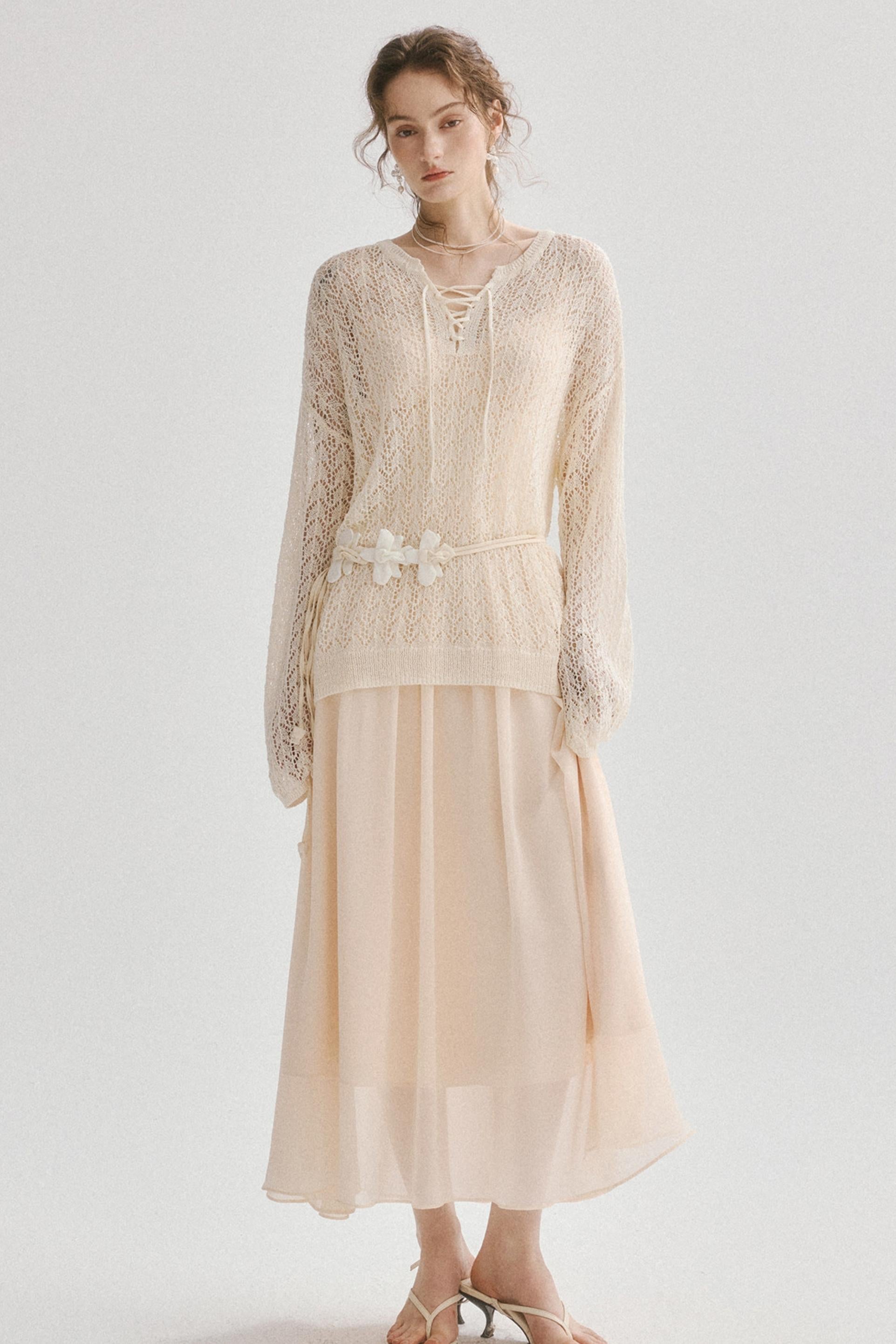 A woman modeling a cream-colored two-piece outfit consisting of a long-sleeve lace top with a lace-up neckline and a matching pleated skirt, styled with a decorative fabric belt and white sandals against a plain light gray background.