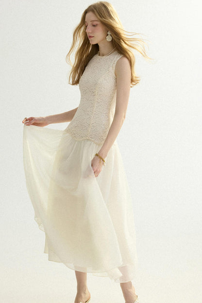 A woman modeling a sleeveless cream-colored dress with a textured lace bodice and a flowing chiffon skirt, set against a plain white studio background.