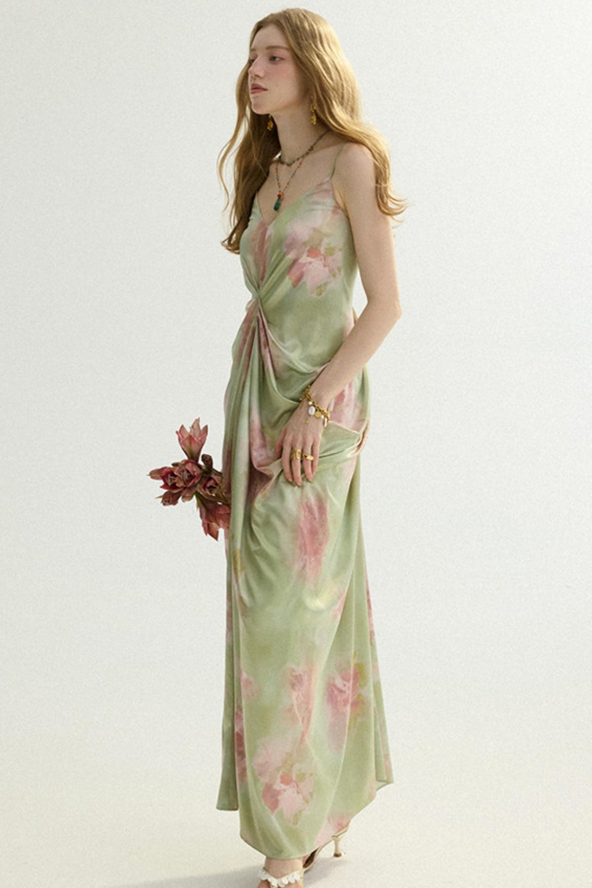 A woman modeling a light sage green maxi dress with a pink floral print, standing against a plain off-white studio background.
