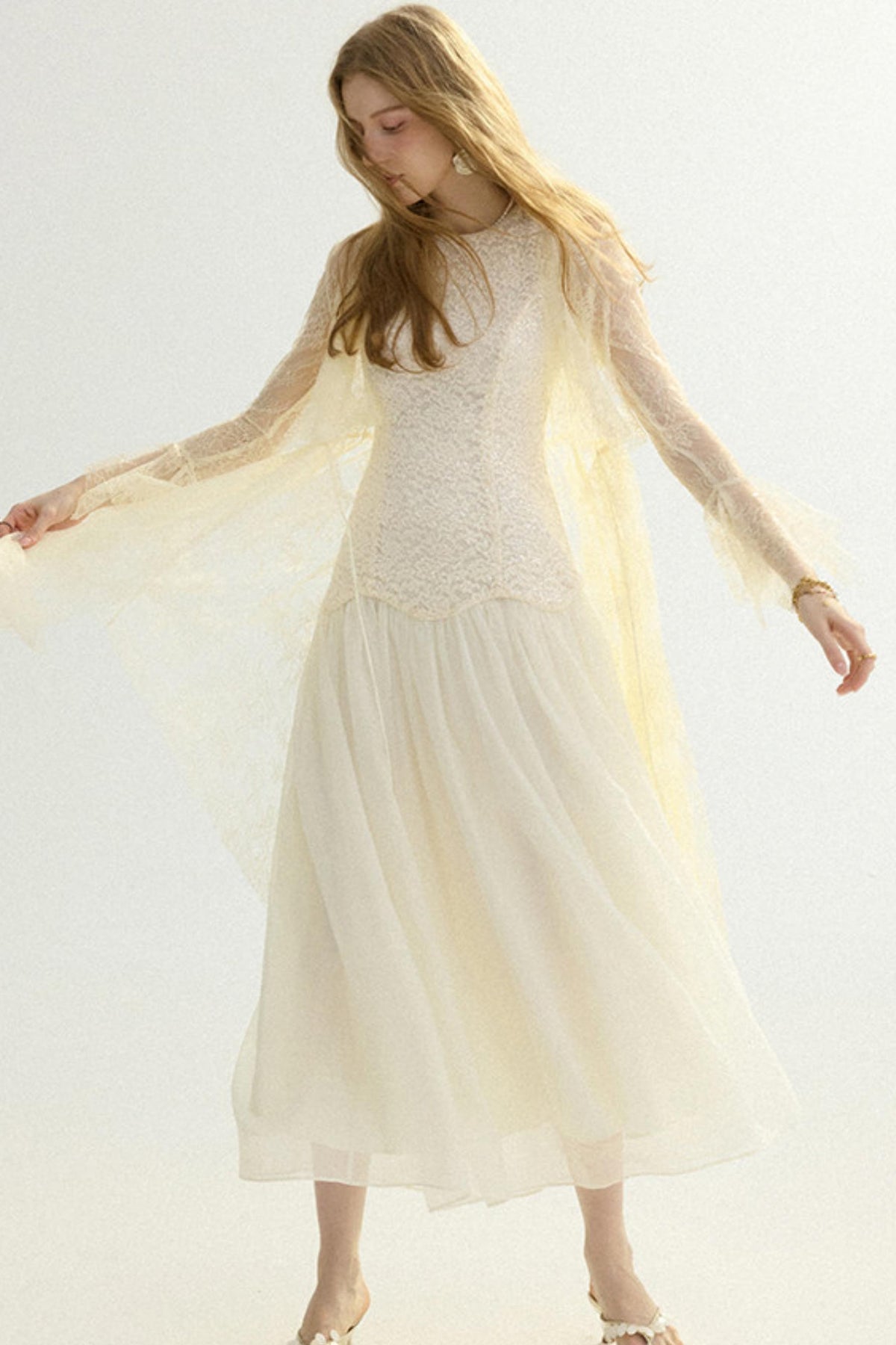 A woman modeling a cream-colored bohemian-style maxi dress with a sheer lace bodice, long flutter sleeves, and a flowing tulle skirt, set against a plain light background.