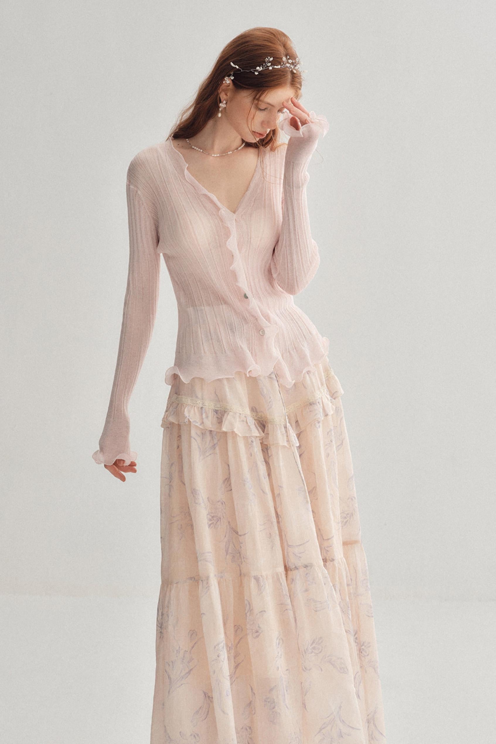 A woman modeling a long-sleeve light pink knit top with a ruffled V-neckline, paired with a matching long skirt featuring a subtle floral pattern, set against a plain light gray background.