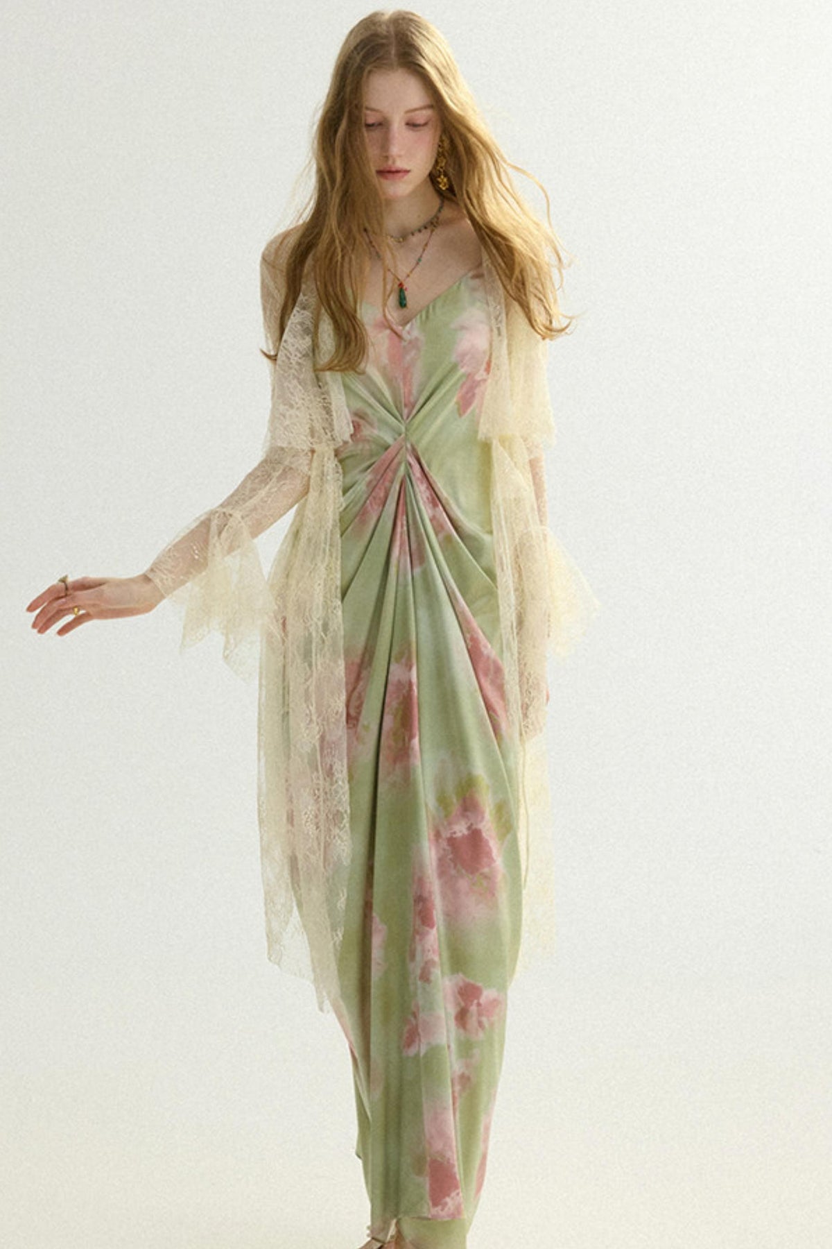 A woman modeling a long, flowing dress with a light sage green base and pink floral print, layered with a sheer cream tulle kimono-style overlay, against a plain light background.