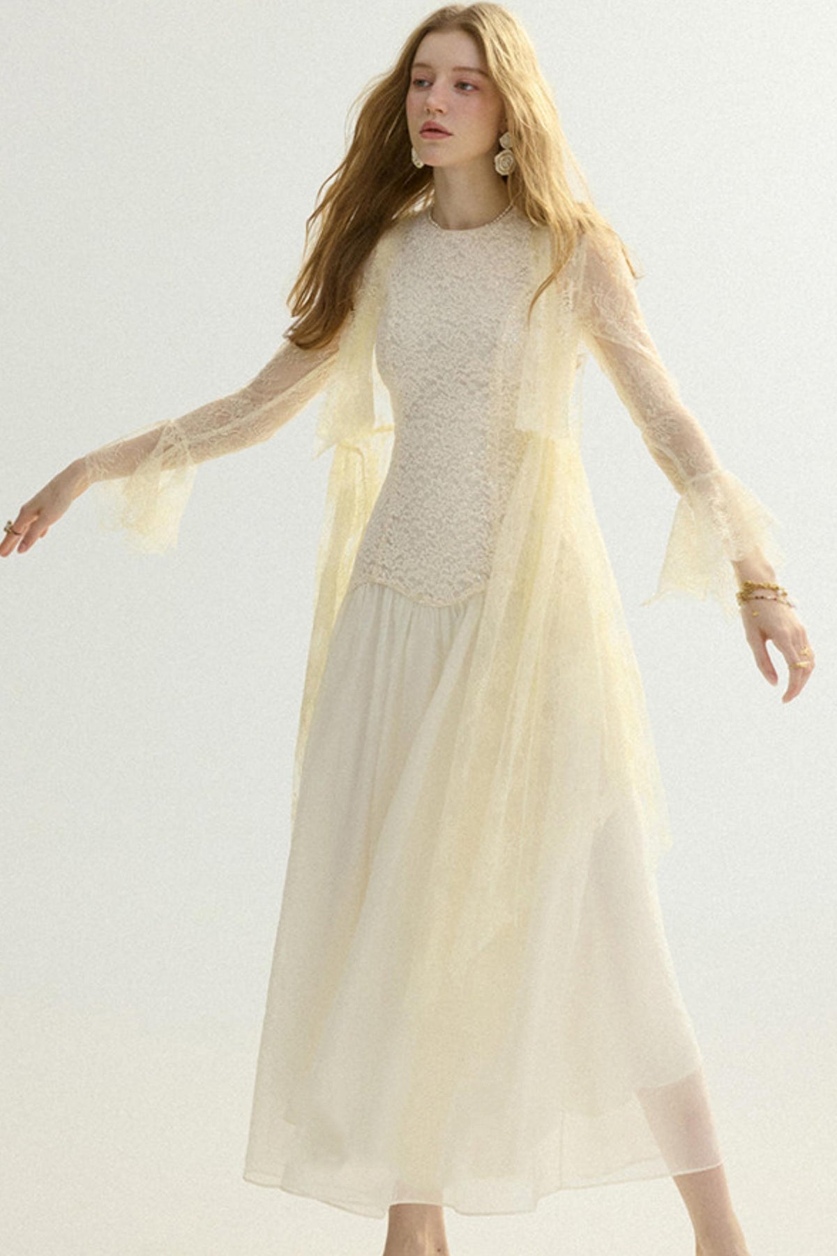 A woman modeling a cream-colored midi dress with a textured lace bodice, sheer long sleeves with ruffled cuffs, and a flowing tulle skirt, set against a plain light background.