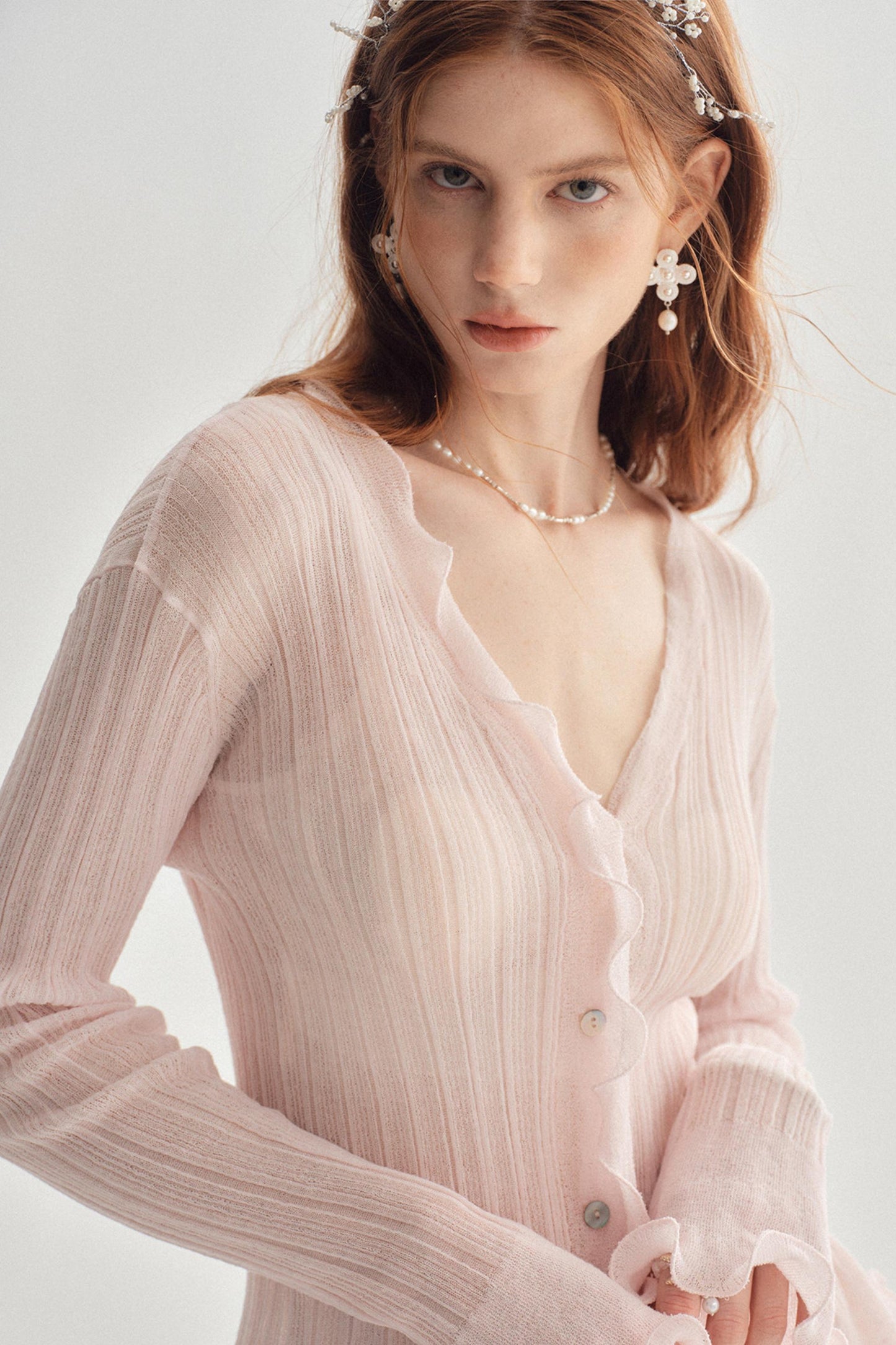 A woman wearing a light pink long-sleeve cardigan with a ruffled V-neck and button front, styled with pearl-like jewelry against a soft light gray background.