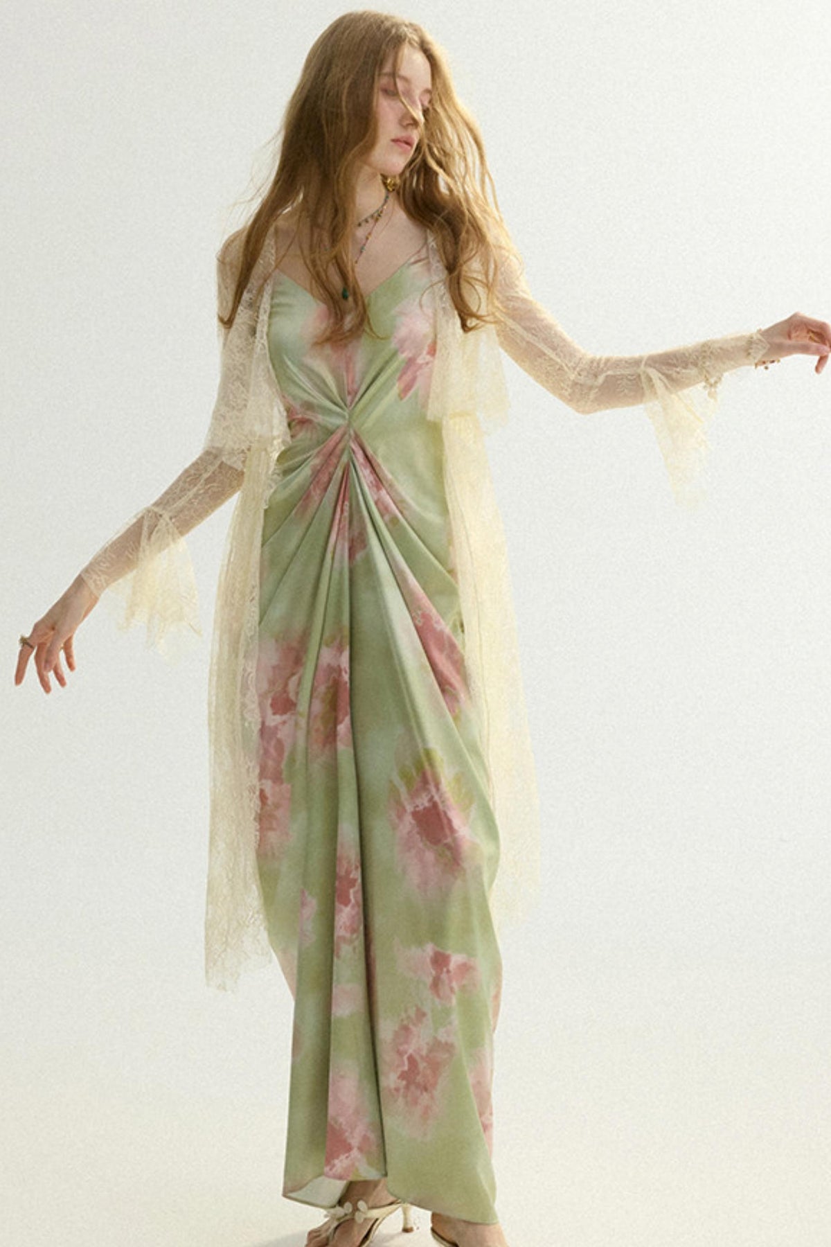 A woman modeling a long sage green maxi dress with pink floral patterns and a sheer cream lace overlay, set against a plain white studio background.