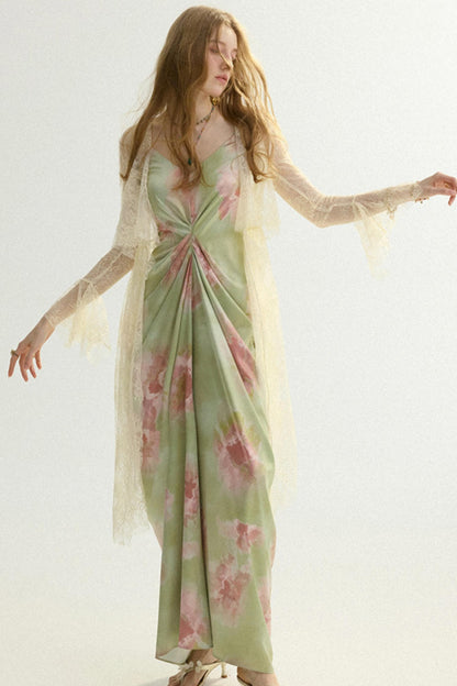 A woman modeling a long sage green maxi dress with pink floral patterns and a sheer cream lace overlay, set against a plain white studio background.