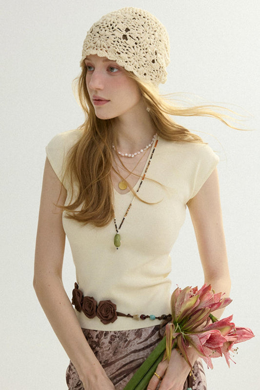 A woman wearing a cream crochet beanie, a cream short-sleeve top, a brown fabric flower belt, and layered necklaces, holding pink flowers against a plain light background.