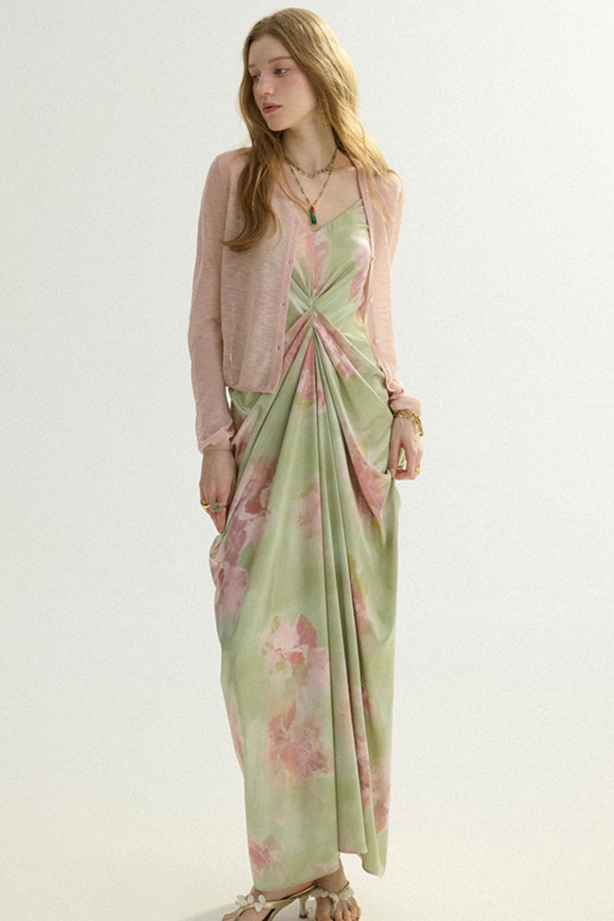 A woman modeling a long-sleeve light pink cardigan layered over a green satin slip dress with a pink floral print and a twisted waist detail, set against a plain light background.