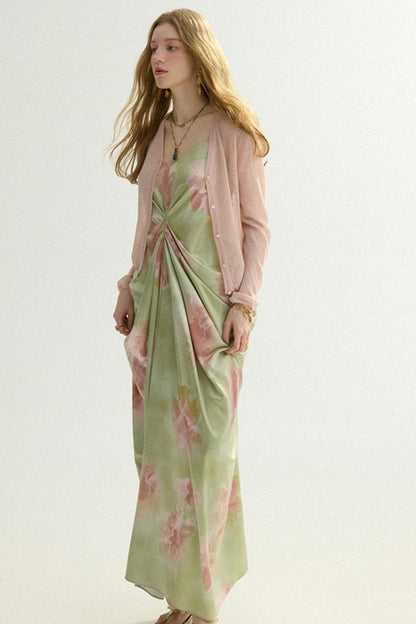 A woman modeling a long light green maxi dress with a pink floral print, styled with a sheer light pink cardigan, against a plain off-white studio background.