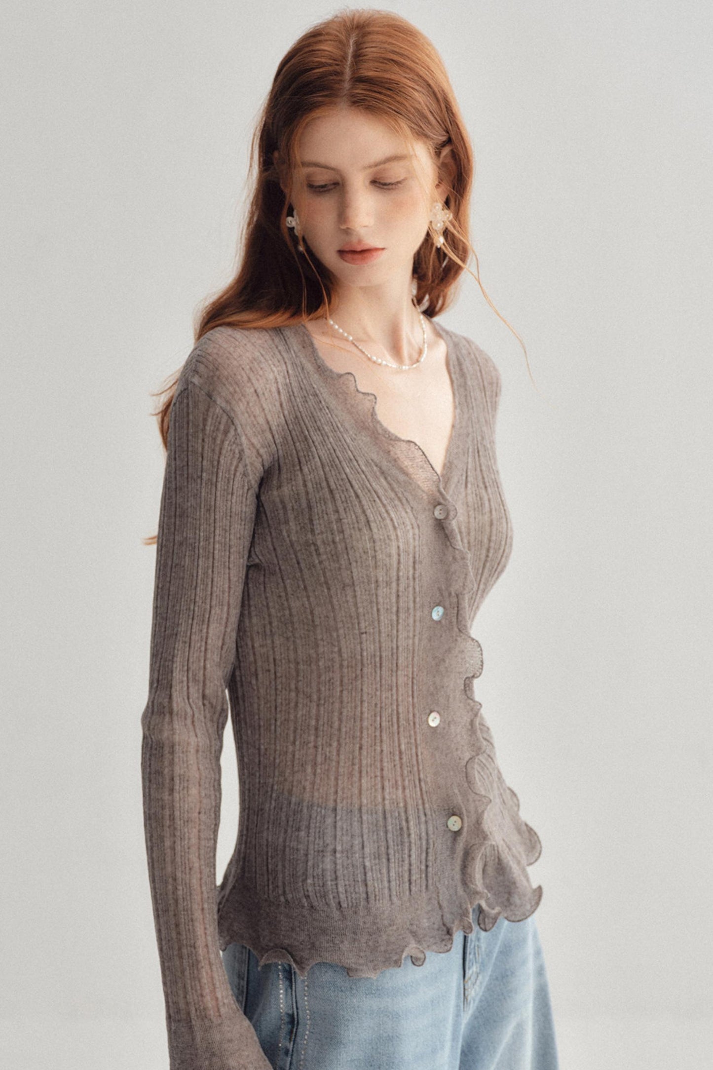 A woman wearing a taupe ribbed knit cardigan with scalloped edges and button details, paired with light blue jeans, photographed against a neutral light gray background.