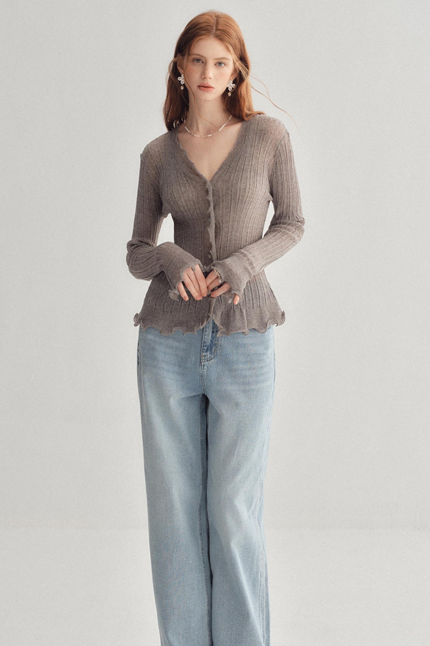 A woman modeling a taupe ribbed knit cardigan with a ruffled hem and button front, paired with light-wash wide-leg jeans, against a plain light gray studio background.