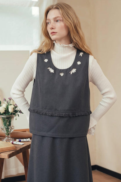 A woman wearing a dark gray sleeveless pinafore-style vest over a cream ribbed turtleneck and matching dark skirt, standing indoors near a table with flowers.