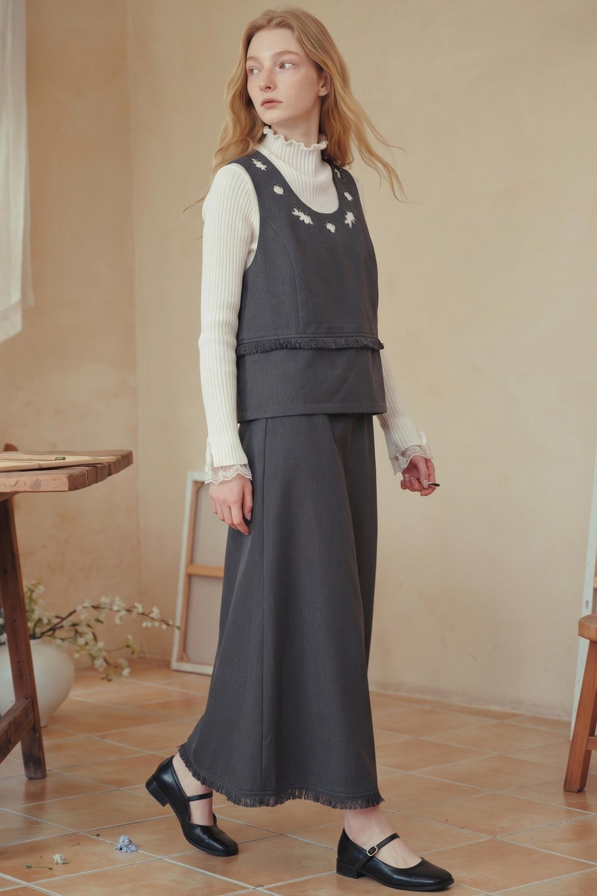 A woman models a dark gray sleeveless vest and matching wide-leg skirt set over a white ribbed turtleneck, paired with black Mary Jane shoes in a warm, rustic indoor setting.