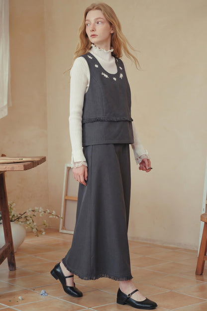A woman models a dark gray sleeveless vest and matching wide-leg skirt set over a white ribbed turtleneck, paired with black Mary Jane shoes in a warm, rustic indoor setting.