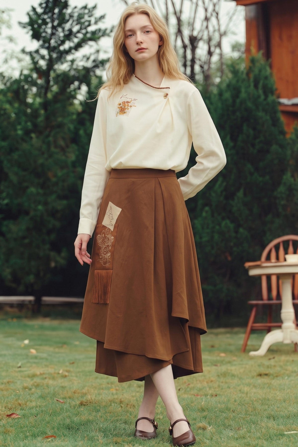 A woman stands outdoors on a grassy lawn wearing a cream long-sleeve blouse with a small embroidered floral detail and a brown tiered midi skirt with patchwork pockets. Trees and a wooden chair are visible in the background.