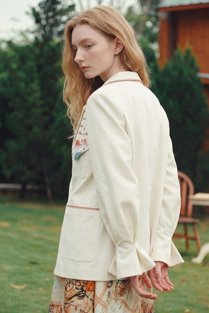 A woman wearing a cream-colored linen jacket with embroidered details and a patterned skirt, standing outdoors in a garden setting.