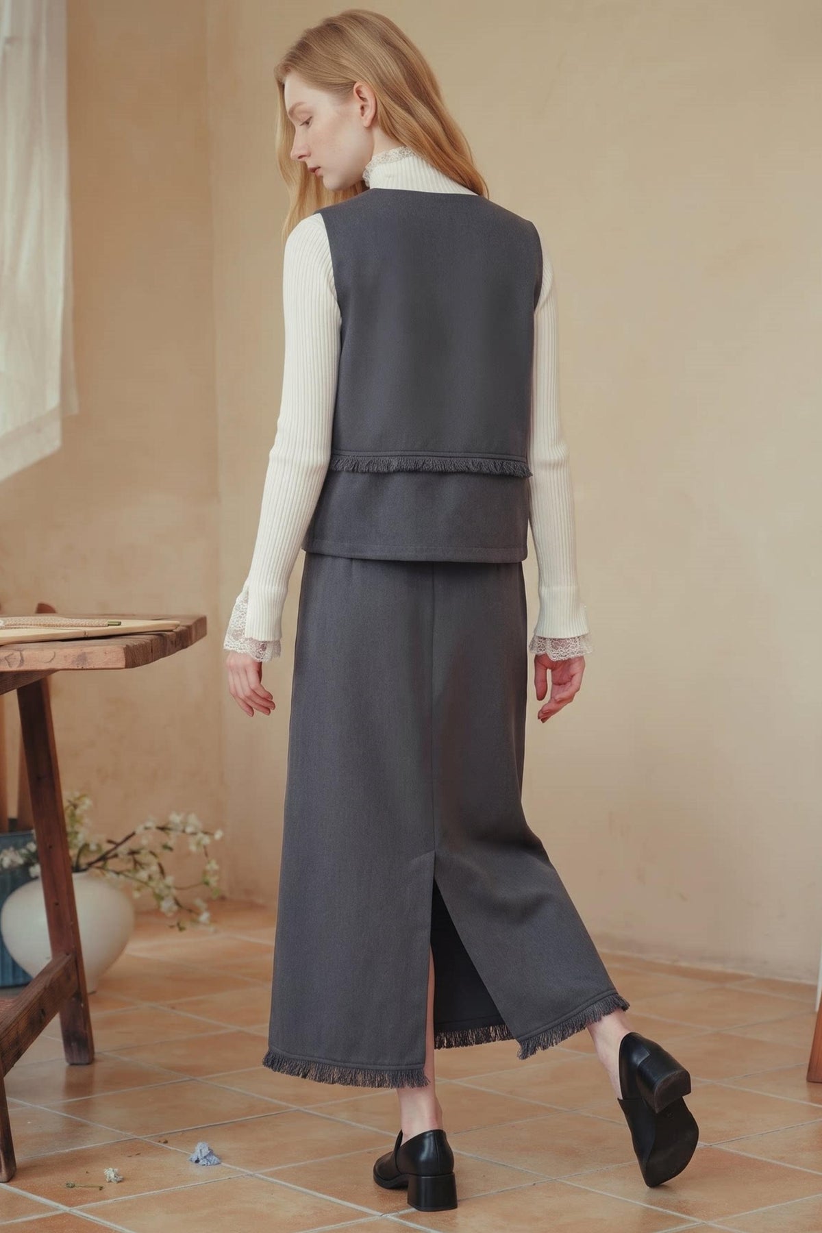 A woman wearing a dark gray sleeveless vest and matching wide-leg skirt with fringe, styled with a cream long-sleeve top and black loafers, standing in a warm beige room.