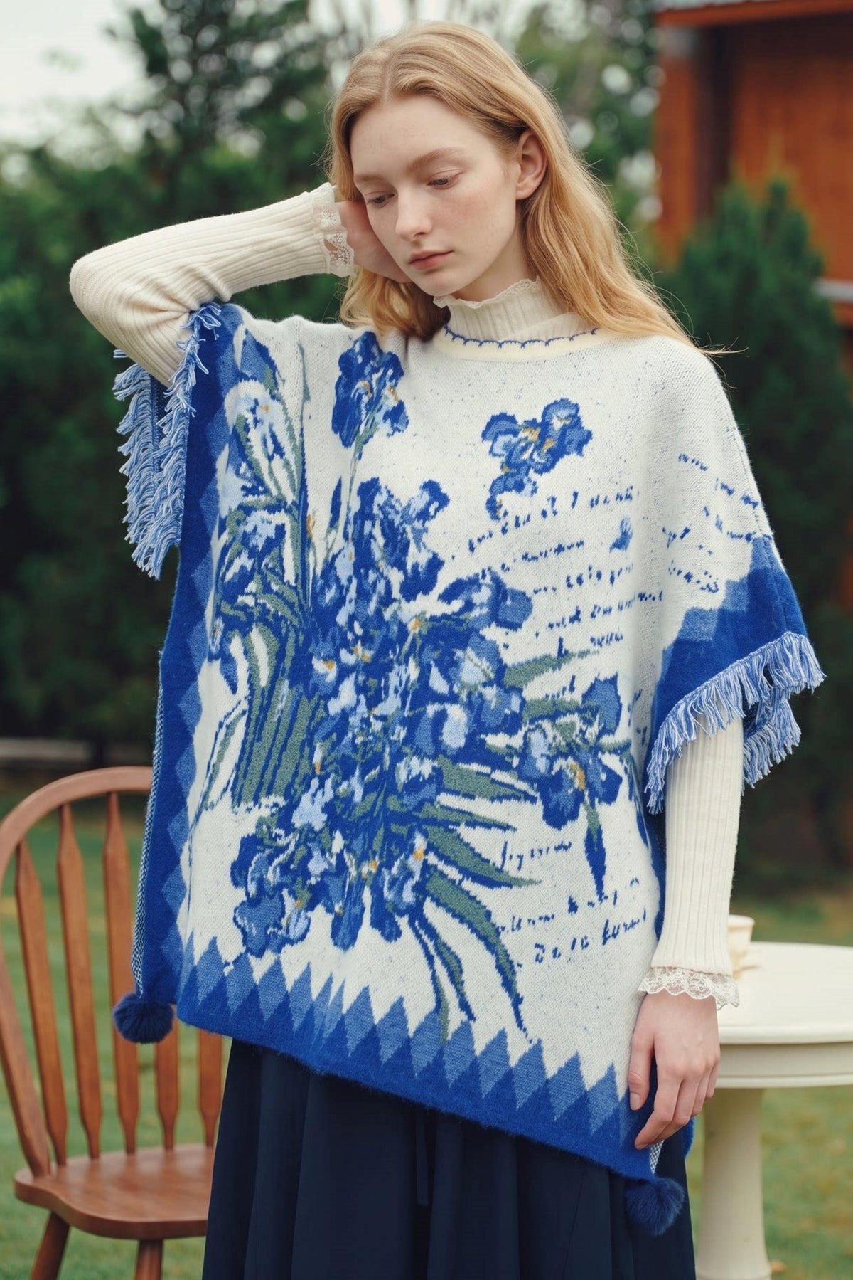A woman wearing a blue and white knit poncho with a floral pattern and fringe, layered over a cream turtleneck and dark skirt, standing outdoors.