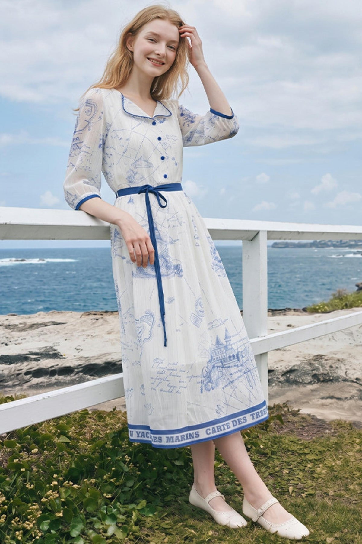 A woman stands outdoors by a white railing overlooking the ocean, wearing a long white dress with blue map-style print and a blue waist tie, paired with white flats.