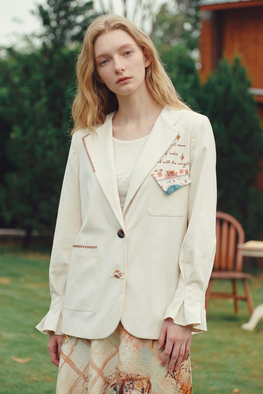A woman standing outdoors in a garden wearing a cream-colored blazer with decorative embroidery and a matching patterned skirt.