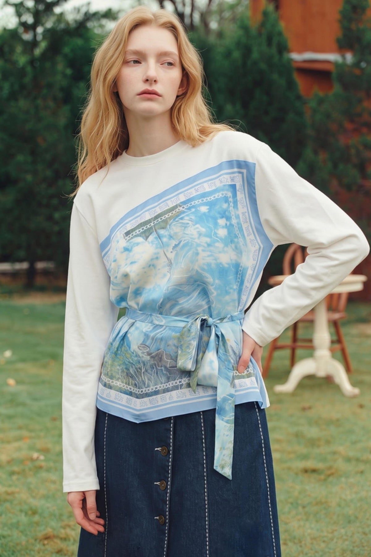 A woman standing outdoors in a grassy area wearing a white long-sleeve top with a large blue and white printed panel across the chest and a matching tied sash, paired with a dark denim skirt.