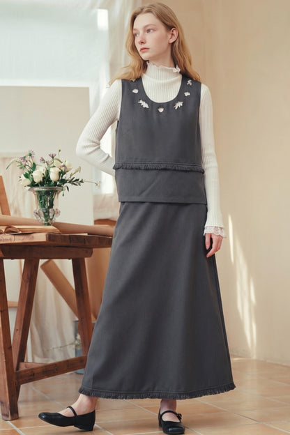 A woman models a dark gray sleeveless vest and matching long skirt set over a white ribbed turtleneck sweater, paired with black Mary Jane shoes in a warm, sunlit room.
