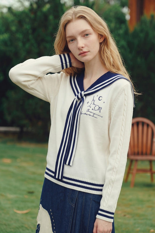 A young woman with long blonde hair stands outdoors in a grassy area wearing a cream-colored knit cardigan with navy blue sailor-style trim and a matching tie, paired with a navy skirt.