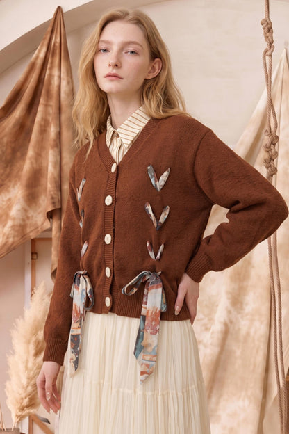 A woman models a brown knit cardigan with decorative tie details and a cream pleated skirt in a warm, rustic studio setting.