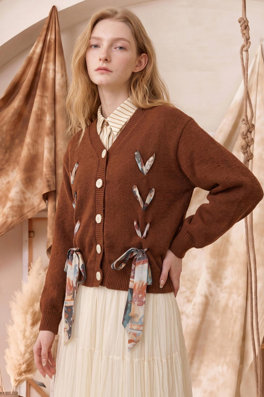 A woman models a brown knit cardigan with decorative tie details and a cream pleated skirt in a warm, rustic studio setting.