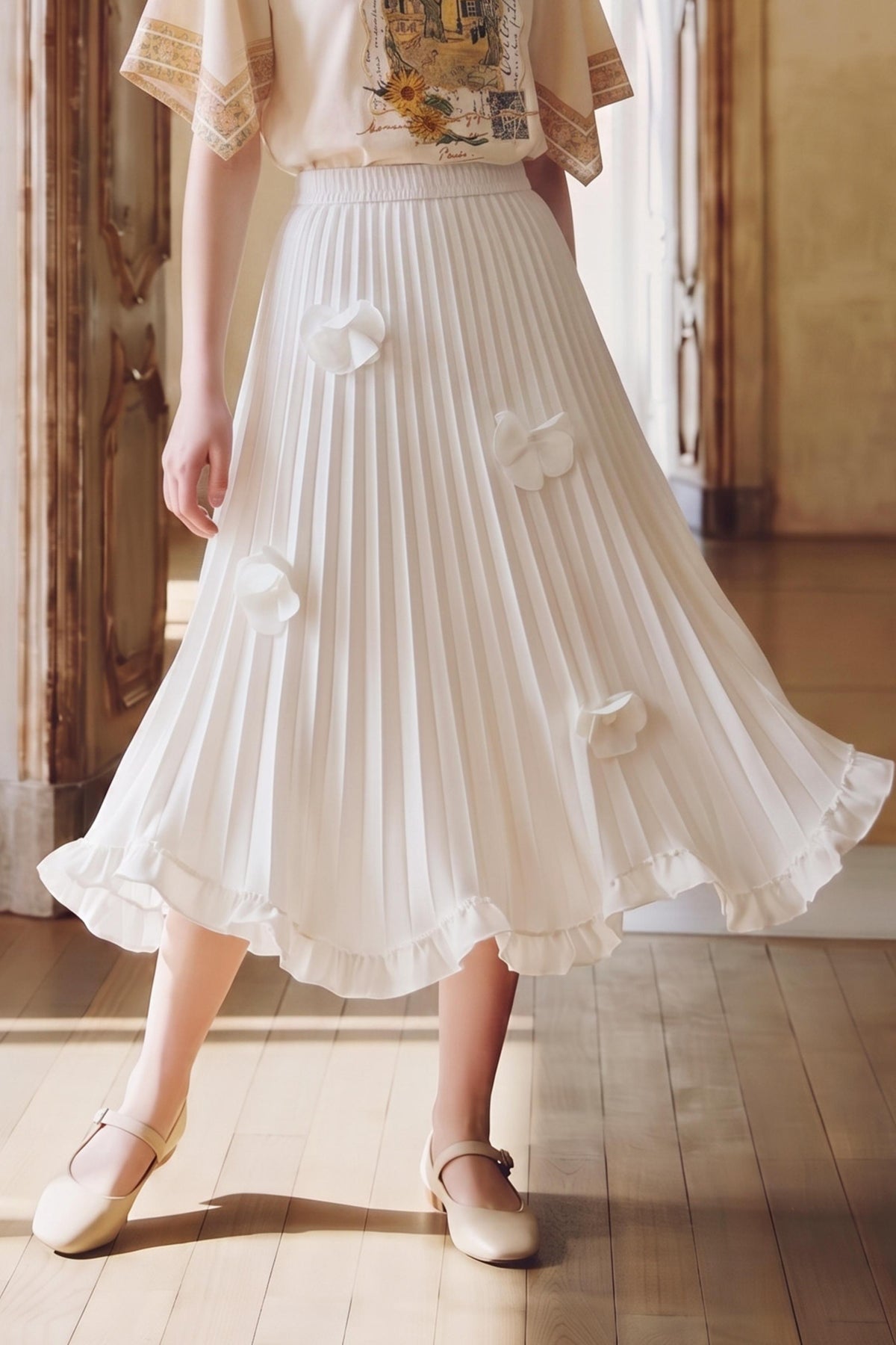 A model wearing a white pleated midi skirt with 3D fabric flower appliqués, paired with a cream blouse and beige Mary Jane shoes, standing in a sunlit room with wooden floors.