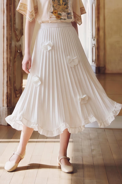 A model wearing a white pleated midi skirt with 3D fabric flower appliqués, paired with a cream blouse and beige Mary Jane shoes, standing in a sunlit room with wooden floors.