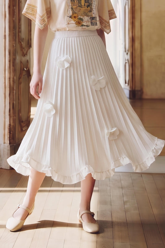 A model wearing a white pleated midi skirt with 3D fabric flower appliqués, paired with a cream blouse and beige Mary Jane shoes, standing in a sunlit room with wooden floors.