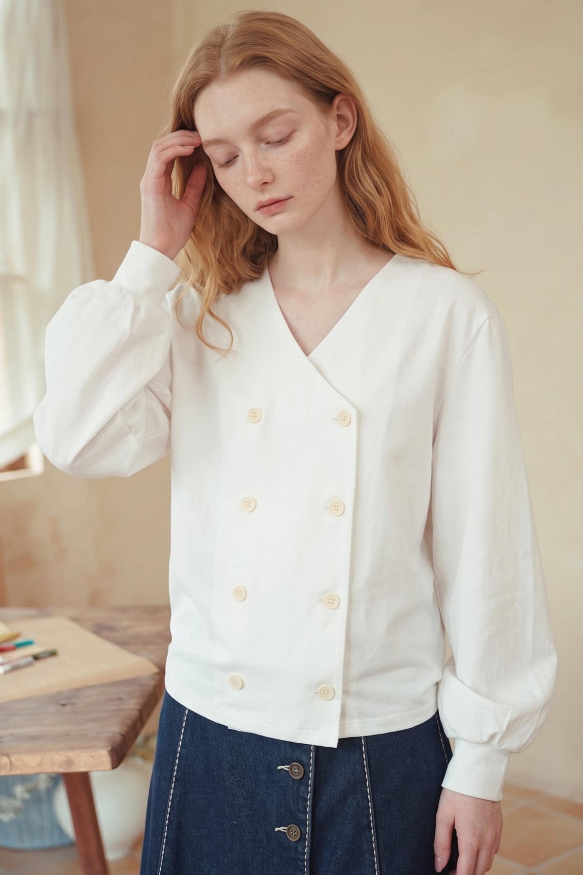 A woman wearing a white long-sleeve wrap-style blouse with cream buttons and dark blue denim skirt, standing in a softly lit indoor setting.
