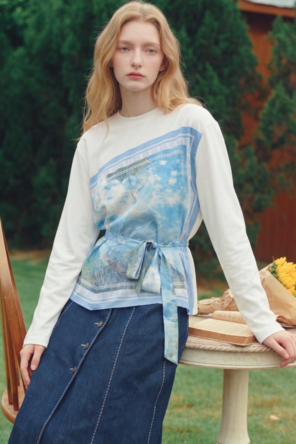 A woman wearing a white long-sleeve top with a blue abstract graphic print and tie waist, paired with a dark blue denim skirt, posed outdoors next to a table.