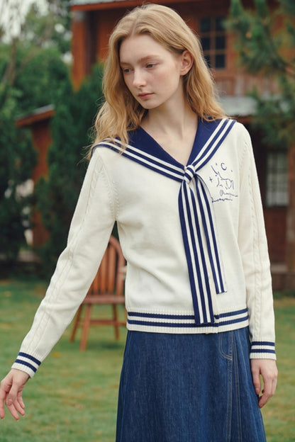 A young woman wearing a cream cable-knit cardigan with a navy blue sailor collar and tie-front stripes, paired with a blue denim skirt, standing outdoors in front of a wooden house.