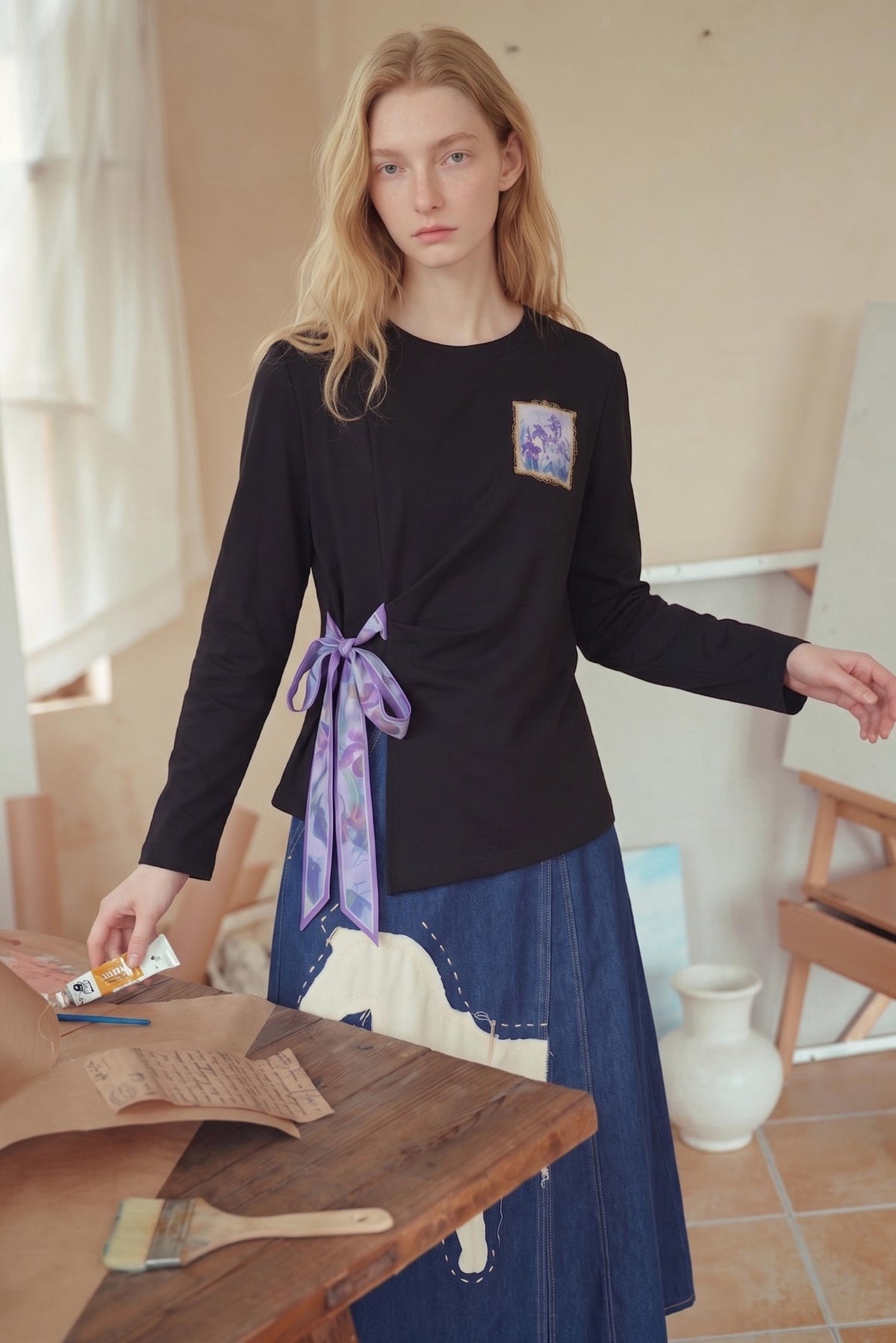 A woman stands in a softly lit studio or room wearing a black long-sleeve top with a small square floral patch on the chest and a purple patterned scarf tied at the waist, paired with a blue skirt. A wooden table with paintbrushes and paper is in the foreground.