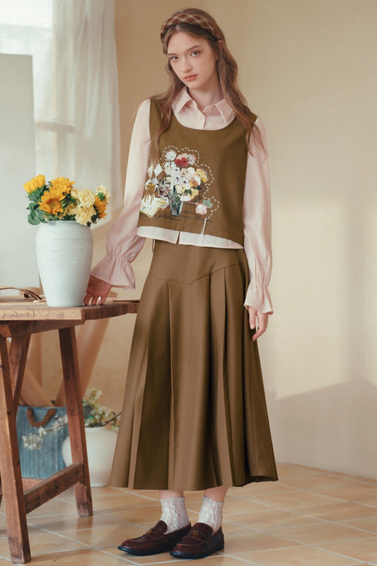 A woman models a brown sleeveless vest with a floral embroidery design over a light pink long-sleeve blouse, paired with a matching pleated brown midi skirt, white socks, and brown loafers in a warm, sunlit room.